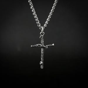 Puede incluir: Un collar con colgante plateado con un diseño de cruz texturizada. La cruz cuelga de una cadena a juego, creando un contraste llamativo sobre el fondo oscuro. El collar es un accesorio religioso.