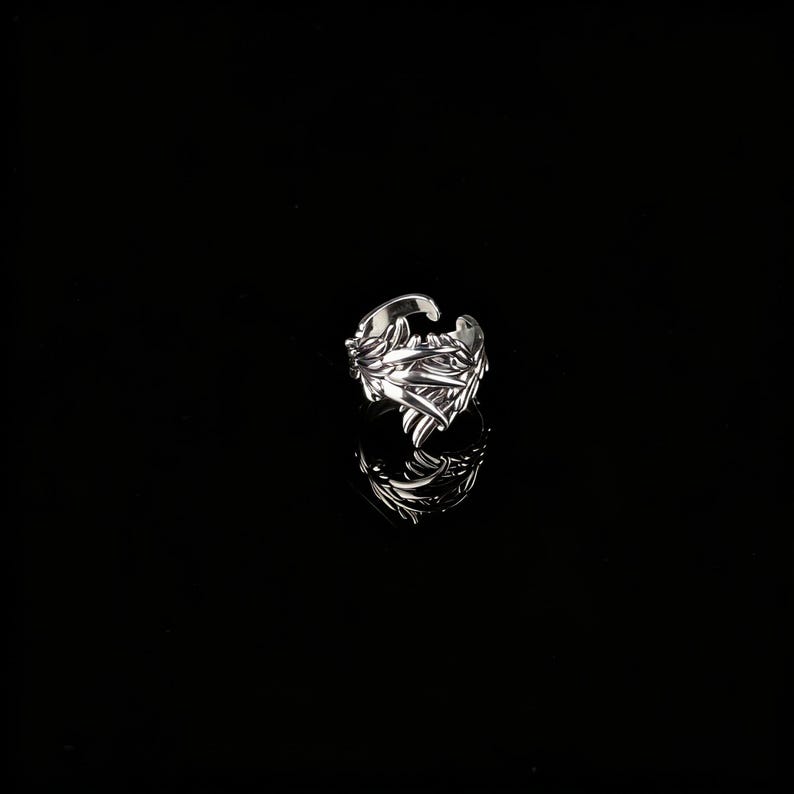 Peut inclure: Une bague argent&eacute;e orn&eacute;e d'un motif de feuilles. La bague pr&eacute;sente un design ouvert et se d&eacute;tache sur un fond noir. Les d&eacute;tails complexes de la bague sont visibles, refl&eacute;tant la lumi&egrave;re.