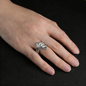 Peut inclure: Une bague en argent avec un motif de feuilles. La bague pr&eacute;sente des motifs de feuilles d&eacute;taill&eacute;s qui entourent le doigt. La bague est pr&eacute;sent&eacute;e sur une main sur fond sombre. La bague est en m&eacute;tal brillant.