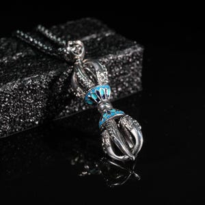 Peut inclure: Un collier pendentif argenté avec un design vajra détaillé, orné d'accents émaillés bleu turquoise. Le pendentif a une structure en forme de griffe en bas et est suspendu à une chaîne délicate. Le fond est sombre et texturé.