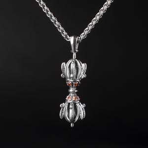 Pode incluir: Um colar com pingente de prata com um design ornamentado de dupla face e detalhes de pedras preciosas vermelhas. O pingente pende de uma corrente trançada sobre um fundo preto. O pingente tem um design simétrico detalhado.