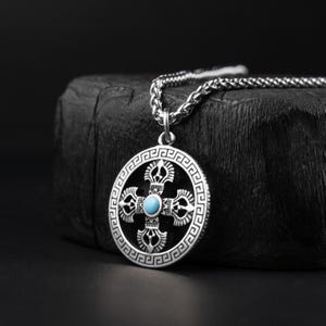 Peut inclure: Collier pendentif argenté avec un motif de croix détaillé et une pierre de couleur turquoise incrustée. Le pendentif circulaire présente une bordure complexe et est suspendu à une chaîne assortie. Le collier est présenté sur un fond sombre.