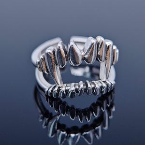 Puede incluir: Un anillo plateado con diseño de colmillos. El anillo presenta una hilera de dientes afilados y puntiagudos, creando una apariencia de vampiro. El anillo se encuentra sobre un fondo oscuro y reflectante, resaltando su brillo metálico.