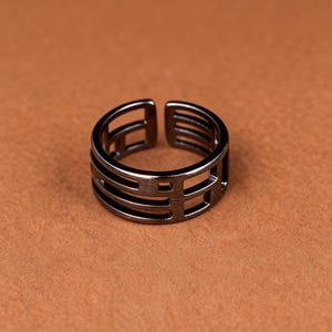 Peut inclure: Une bague réglable gris foncé avec un design géométrique. La bague présente une série de barres horizontales et verticales, créant une esthétique ouverte et moderne. La bague est posée sur un fond marron.