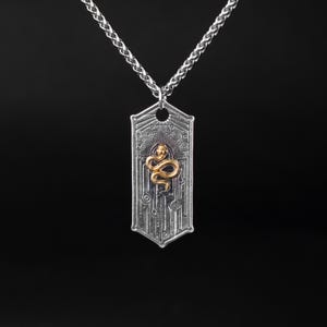 May include: Silver-toned pendant necklace with a gold-colored snake design. The rectangular pendant features intricate etched patterns and a small hole at the top for the chain. The snake is coiled in the center of the pendant. The chain is a braided style.