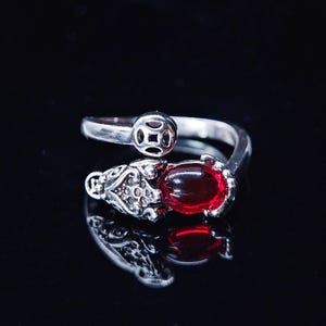 May include: A silver-toned ring with an adjustable design. The ring features a large, oval, deep red gemstone and ornate detailing on the band. The ring's design includes a coin-like element and a decorative setting around the gemstone.