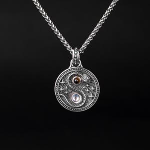 Peut inclure: Un collier pendentif argenté avec un pendentif circulaire représentant un design Yin Yang. Le pendentif a une bordure texturée et un design détaillé avec une pierre marron et une pierre blanche. La chaîne est un design à maillons.
