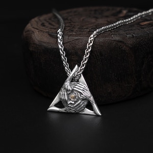 May include: Silver-toned pendant necklace featuring a triangular pendant with a detailed design of hands encircling a central element. The necklace has a chain with a textured pattern, and the pendant has a small gold-colored accent.