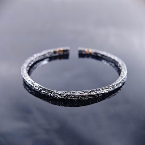 May include: A silver-coloured, open-ended bracelet with a textured surface. The bracelet has a dark, almost black, appearance with small gold-coloured accents near the open ends. The bracelet is displayed on a reflective surface.