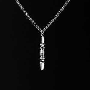 Peut inclure: Un collier pendentif argenté avec un pendentif vertical détaillé. Le pendentif présente une section centrale ornée et des extrémités pointues. La chaîne est de style tressé, sur fond noir uni.