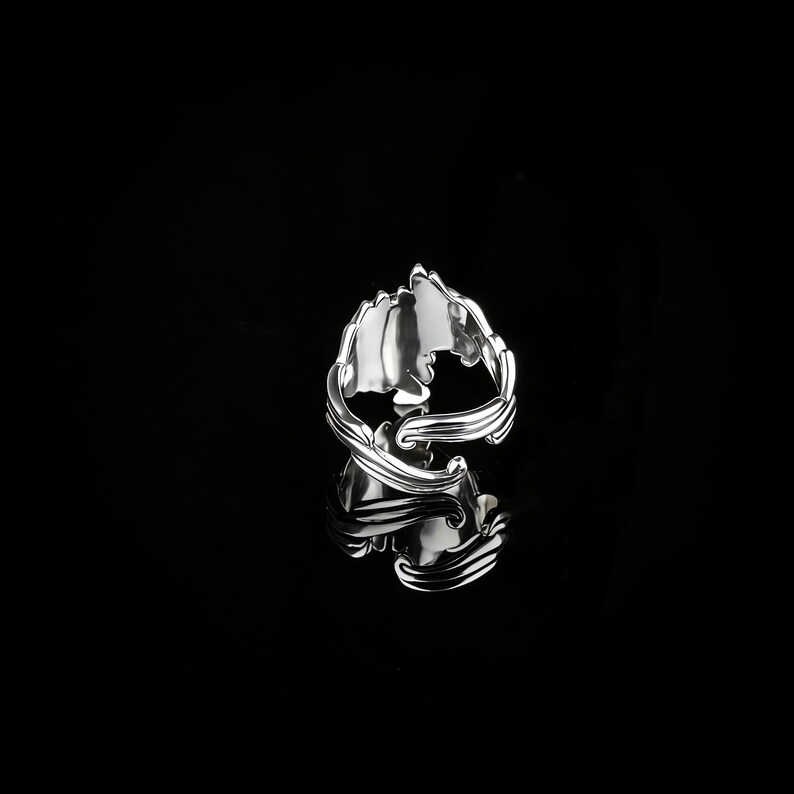 Peut inclure: Une bague en argent avec un design orn&eacute; et tourbillonnant. La bague pr&eacute;sente une forme centrale en forme de feuille et des d&eacute;tails complexes. La bague est pr&eacute;sent&eacute;e sur une surface noire r&eacute;fl&eacute;chissante.
