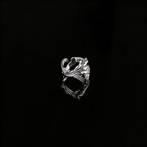 Peut inclure: Une bague en argent avec un design orn&eacute; et fluide. La bague pr&eacute;sente une s&eacute;rie de lignes courbes et de formes en forme de feuilles, cr&eacute;ant un aspect textur&eacute; et d&eacute;taill&eacute;. La bague est pos&eacute;e sur un fond noir, soulignant sa surface r&eacute;fl&eacute;chissante.