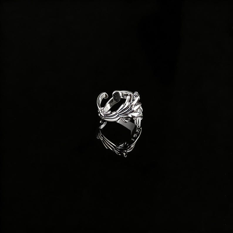 Peut inclure: Une bague en argent avec un design orn&eacute; et fluide. La bague pr&eacute;sente une s&eacute;rie de lignes courbes et de formes en forme de feuilles, cr&eacute;ant un aspect textur&eacute; et d&eacute;taill&eacute;. La bague est pos&eacute;e sur un fond noir, soulignant sa surface r&eacute;fl&eacute;chissante.