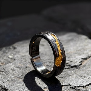 Op de afbeelding: Een zilveren ring met een getextureerde, donkergrijze band en een helder gouden inleg. De ring heeft een verstelbare opening. De ring wordt gepresenteerd op een grijze, getextureerde ondergrond.