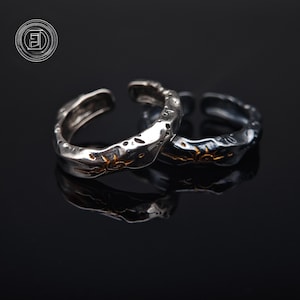 Może przedstawiać: Dwa regulowane pierścionki w kolorze srebrnym o teksturowanym, organicznym wzorze. Każdy pierścionek ma złoty wzór słońca. Pierścionki są prezentowane na odblaskowej czarnej powierzchni, podkreślając ich metaliczny połysk.