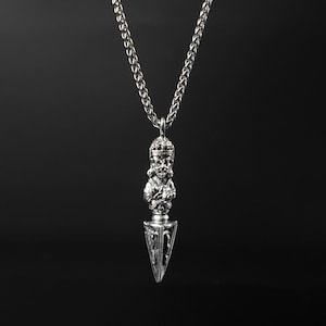 Puede incluir: Collar con colgante plateado con una figura detallada y un diseño de punta de flecha estilizada. El colgante cuelga de una cadena con un patrón tejido. El fondo es negro sólido.
