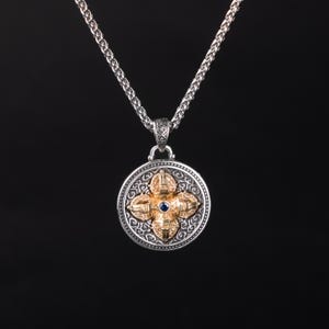 May include: A silver pendant necklace with a detailed, circular pendant. The pendant features a gold-colored, four-petal design with a blue gemstone center. The necklace chain is also silver and has a textured appearance.