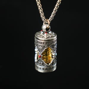 May include: A silver pendant necklace with an ornate cylindrical charm. The charm features intricate patterns, a gold-colored center, and is adorned with red and blue gemstones. The chain is a silver braided style, and the background is black.