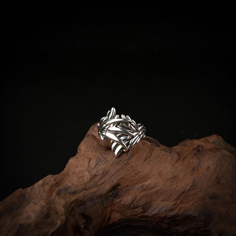 Peut inclure: Une bague en argent avec un motif de feuilles d&eacute;taill&eacute;. La bague est faite d'un m&eacute;tal brillant et pr&eacute;sente de multiples formes de feuilles qui se chevauchent. La bague est pr&eacute;sent&eacute;e sur un morceau de bois sur fond sombre.