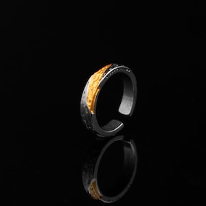 May include: An adjustable silver ring featuring a gold-coloured textured inlay. The ring has a dark, hammered texture on the outer band and a reflective surface. The open-ended ring is set against a black background.