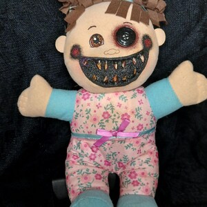 May include: A spooky doll with brown pigtails, a button eye, and a stitched grin filled with sharp teeth. The doll is dressed in a pink floral jumpsuit with blue sleeves and feet, and a pink bow. The doll's face has dark painted accents.