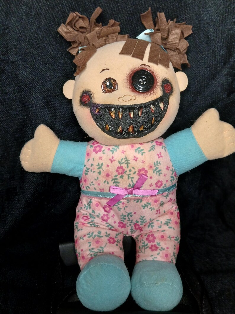 May include: A spooky doll with brown pigtails, a button eye, and a stitched grin filled with sharp teeth. The doll is dressed in a pink floral jumpsuit with blue sleeves and feet, and a pink bow. The doll's face has dark painted accents.