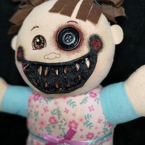 May include: A handmade doll with a button eye and a stitched, menacing grin. The doll's face is painted with dark accents, and it wears a floral print dress with a pink bow. Brown fabric is used for the doll's hair.