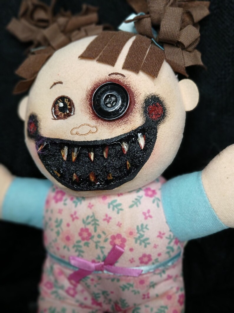 May include: A handmade doll with a button eye and a stitched, menacing grin. The doll's face is painted with dark accents, and it wears a floral print dress with a pink bow. Brown fabric is used for the doll's hair.