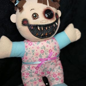 May include: A handmade plush doll with a disturbing appearance. The doll has a button eye and a gaping, tooth-filled mouth. It wears a pink floral patterned outfit with blue accents and a pink bow. The doll's hair is styled in brown pigtails.