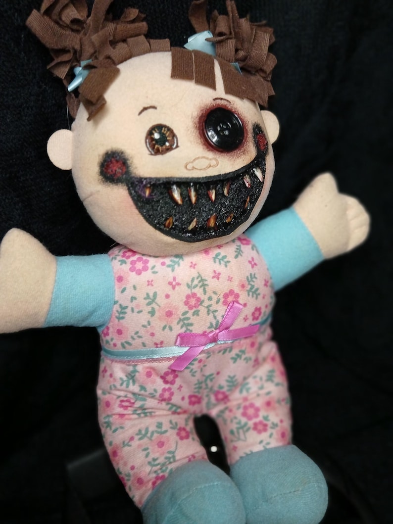 May include: A handmade plush doll with a disturbing appearance. The doll has a button eye and a gaping, tooth-filled mouth. It wears a pink floral patterned outfit with blue accents and a pink bow. The doll's hair is styled in brown pigtails.