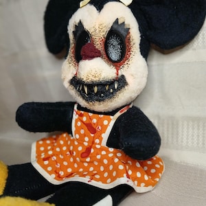 May include: A plush doll with a disturbing, horror-themed design. The doll features black ears, a white face with a grotesque expression, and an orange polka-dot apron with red splatters. The doll's eyes are black, and it has sharp teeth.