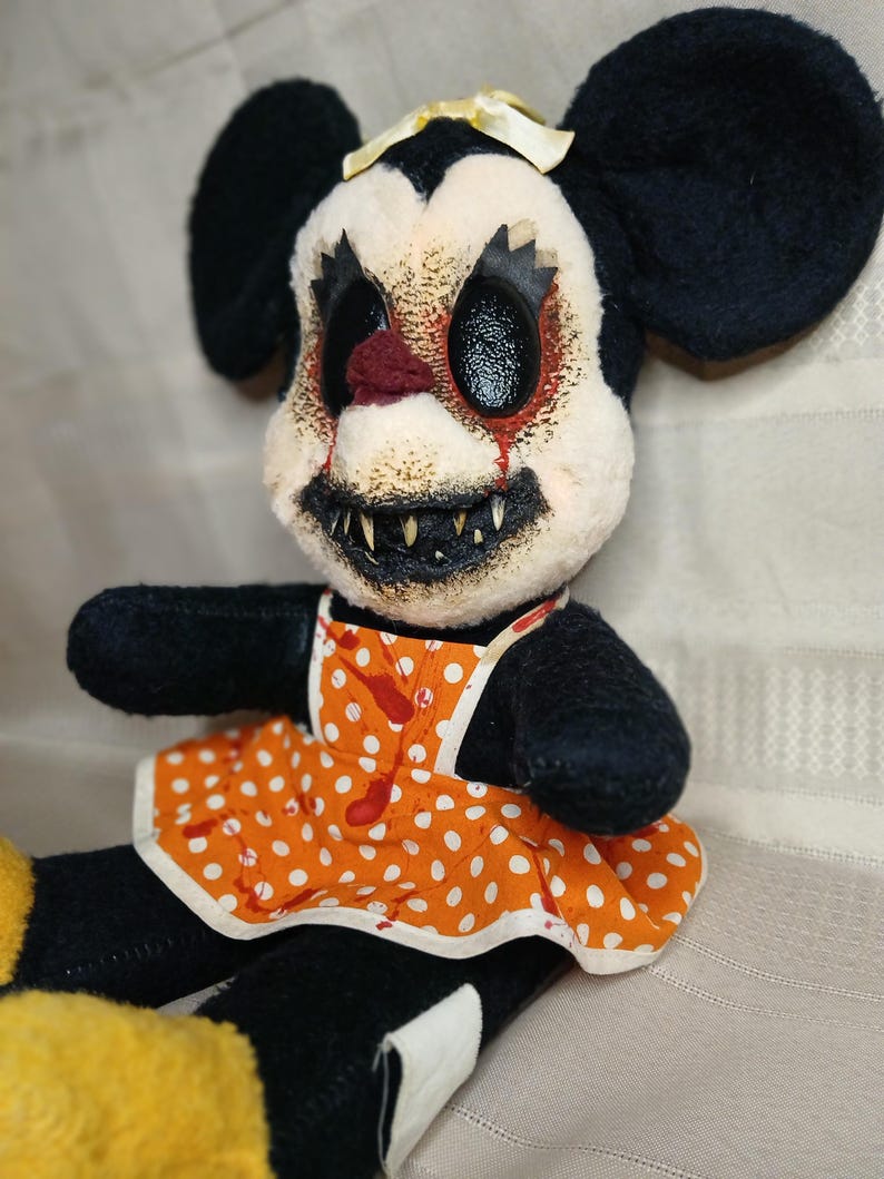 May include: A plush doll with a disturbing, horror-themed design. The doll features black ears, a white face with a grotesque expression, and an orange polka-dot apron with red splatters. The doll's eyes are black, and it has sharp teeth.