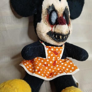 May include: A creepy, handmade plush doll with black ears, a yellow bow, and a sinister expression. The doll wears an orange polka-dot apron with red splatters. Yellow shoes complete the unsettling look.