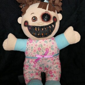 May include: A handmade plush doll with a creepy, horror-themed design. The doll has brown hair, a button eye, and a stitched, toothy grin. It wears a floral print outfit with a pink bow and blue accents. The doll's arms are outstretched.
