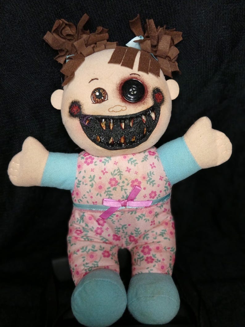 May include: A handmade plush doll with a creepy, horror-themed design. The doll has brown hair, a button eye, and a stitched, toothy grin. It wears a floral print outfit with a pink bow and blue accents. The doll's arms are outstretched.