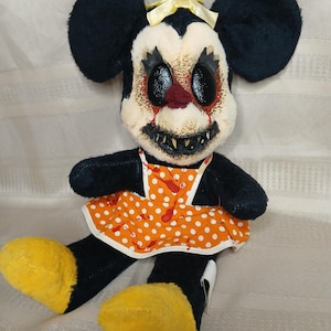 May include: A plush toy of a mouse with black fur, large ears, and yellow feet. The mouse has a white face with a disturbing expression, red accents, and sharp teeth. It wears an orange dress with white polka dots and a yellow bow.