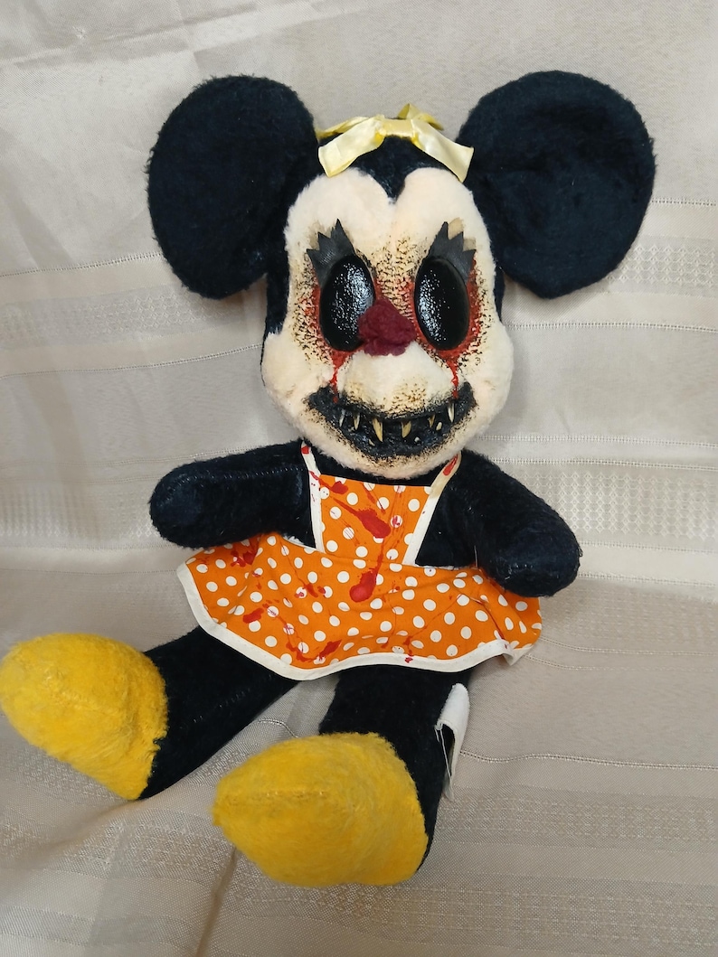 May include: A plush toy of a mouse with black fur, large ears, and yellow feet. The mouse has a white face with a disturbing expression, red accents, and sharp teeth. It wears an orange dress with white polka dots and a yellow bow.