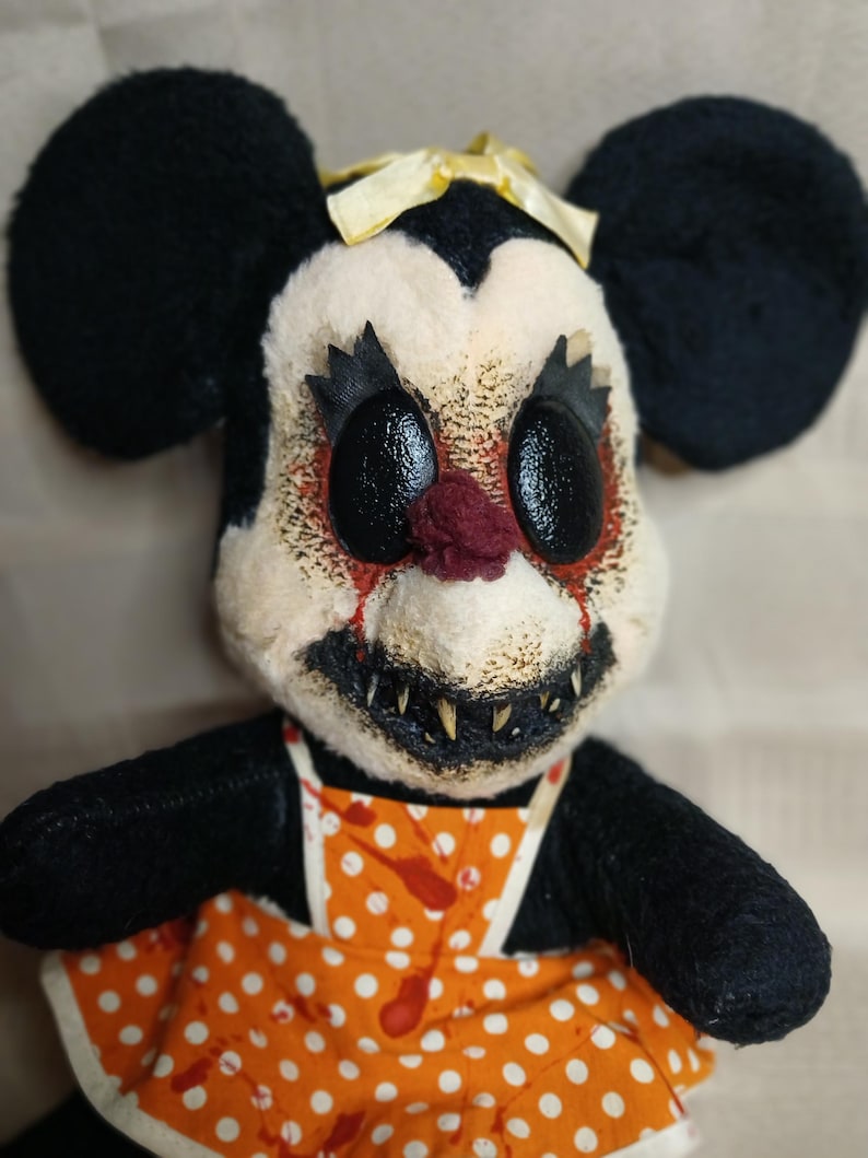 May include: A creepy plush doll with black ears, a yellow bow, and a disturbing face. The doll has large black eyes, a red nose, and a stitched-on mouth with sharp teeth. It wears an orange apron with white polka dots and red blood-like stains.
