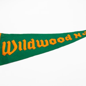 May include: Green felt pennant with orange lettering that reads "Wildwood NJ". The pennant has an orange border and orange tassels. The pennant is a classic souvenir.