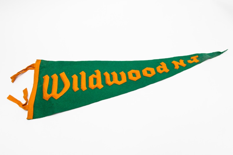 May include: Green felt pennant with orange lettering that reads "Wildwood NJ". The pennant has an orange border and orange tassels. The pennant is a classic souvenir.