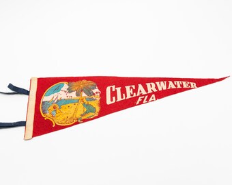 May include: Red felt pennant with the words "CLEARWATER FLA" in white. An illustration of a beach scene with palm trees and people is on the left side. Blue ribbons are attached to the pennant.