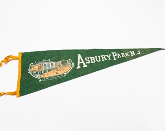 May include: Green felt pennant with gold trim and tassels. The pennant features the words "Asbury Park N.J." in white, along with an illustration of a building and the words "New Casino."