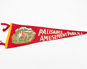 May include: Red felt pennant with yellow trim and red ribbons. The pennant features an illustration of a person on a horse and the text "PALISADES AMUSEMENT PARK, N.J." in white lettering. A vintage souvenir.