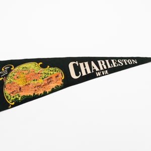 May include: A black felt pennant featuring the text CHARLESTON W.VA. in large cream-colored letters. To the left, a colorful graphic depicts a domed building in orange and yellow tones surrounded by green trees, with the text Visit West Virginia.