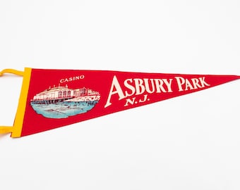 May include: Red pennant with yellow trim featuring the words "Asbury Park N.J." in white script. A detailed illustration of a building labeled "Casino" is also on the pennant. The pennant is a souvenir.