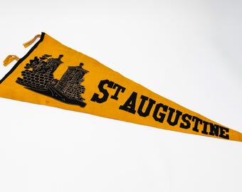 May include: A vintage pennant in a triangular shape, featuring the text "ST AUGUSTINE" in bold black letters. The pennant is yellow with a black border and a black illustration of a building. The pennant has yellow tassels.