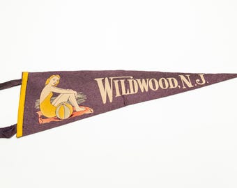 May include: A vintage pennant in a triangular shape, featuring the text "Wildwood, N.J." in cream-colored letters. The pennant has a purple background with an illustration of a woman in a yellow swimsuit sitting on a beach towel with a beach ball.