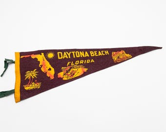 May include: Maroon pennant with gold lettering and illustrations, featuring "DAYTONA BEACH FLORIDA". The pennant has a yellow border and green ties. Illustrations include a map of Florida, a sun, palm trees, and a race car.