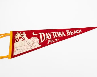 May include: A red felt pennant with the words "Daytona Beach FLA." in white script. The pennant features a beach scene with a palm tree, a woman, and an umbrella. Yellow ribbon is attached to the pennant.