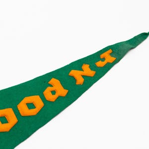 May include: Green felt pennant with orange block letters spelling "WOOD NX". The pennant is a classic shape, with a pointed end. The background is plain white.
